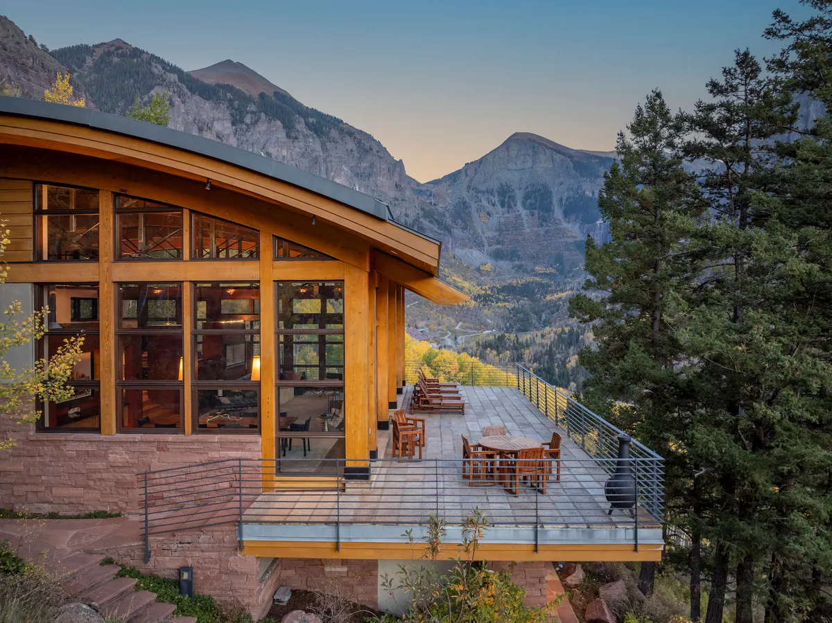 A modern outdoor terrace with a dining table and comfortable seating overlooks majestic mountains. The area is designed for alfresco dining and lounging, with large glass windows offering a seamless indoor-outdoor connection and inviting gatherings under the sky.