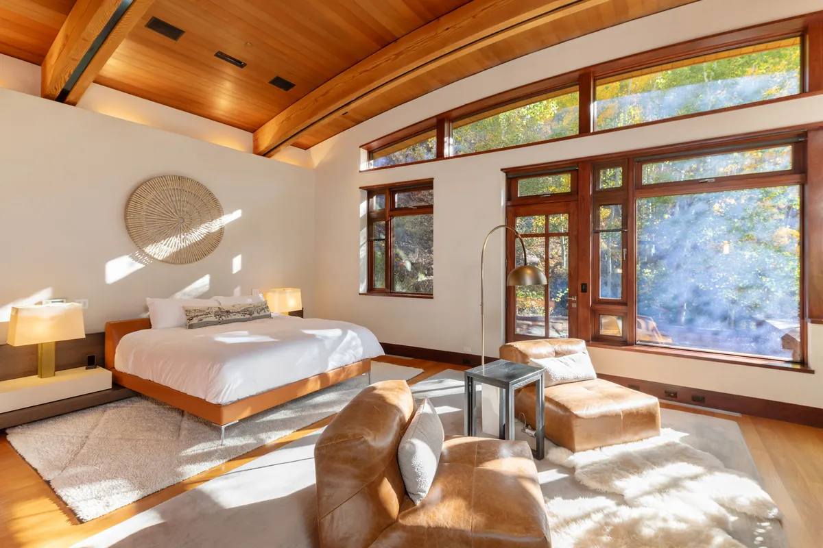 A tranquil bedroom with large windows offering an expansive view of the outdoors, a plush bed with luxury linens, wooden ceiling, lounge chairs, and elegant wall art. The natural light creates a serene, peaceful atmosphere ideal for unwinding.