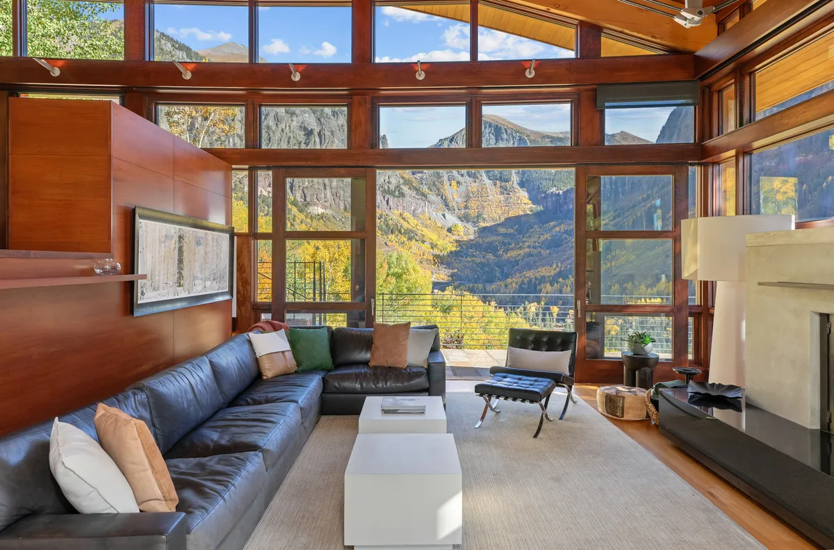 A tranquil living room featuring a large sectional sofa positioned to take full advantage of floor-to-ceiling windows framing a dramatic mountain view. A sleek, modern coffee table anchors the space, while a stylish fireplace offers warmth and ambiance. The room is filled with natural light and has an inviting, open layout perfect for group gatherings or quiet relaxation.