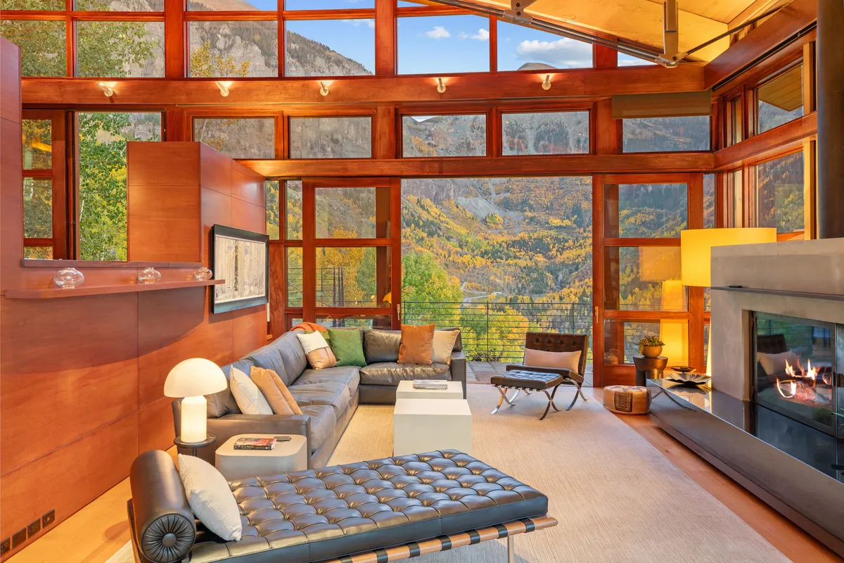 A contemporary, cozy living area with expansive windows that frame the mountains. The room features modern furniture, a welcoming fireplace, and a seating area designed for both intimate gatherings and solo moments of relaxation. Warm, natural light fills the space, emphasizing the home's connection to its surroundings.