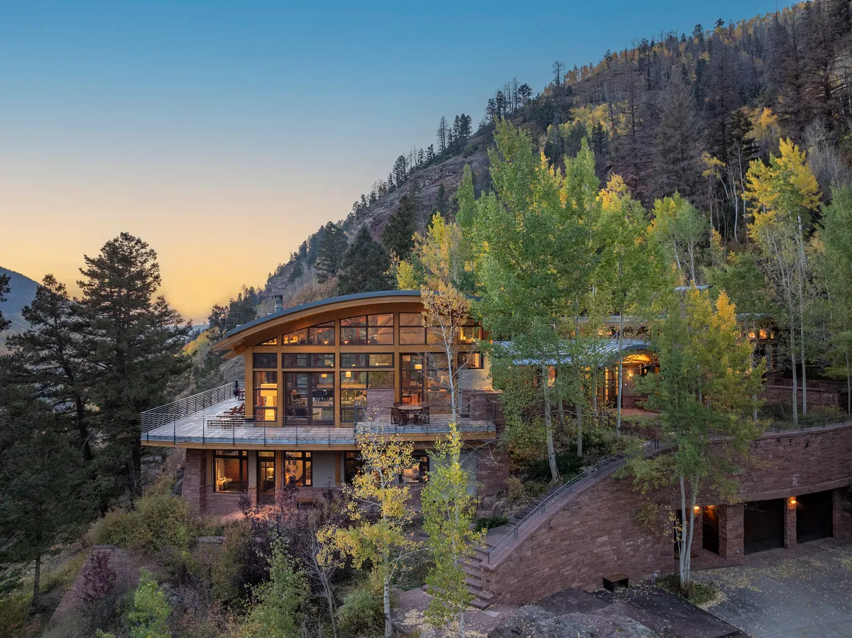 The image showcases a modern architectural home perched on a hillside, surrounded by lush trees and the natural beauty of Telluride. The house features expansive windows and a spacious deck, set against a dazzling sunset sky. Outdoor lounge furniture invites relaxation, while hints of a fire pit and nature trails offer glimpses of activities guests can enjoy.