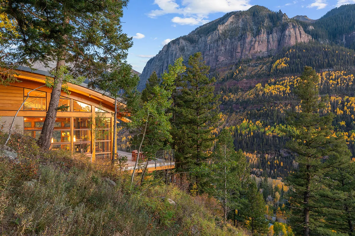 This image showcases the home's expansive exterior, set against a backdrop of mountains and surrounded by lush trees. Large windows invite natural light into the house, and a deck area offers a space for outdoor relaxation or family gatherings. The setting feels tranquil but accessible, hinting at both seclusion and proximity to local parks and attractions.