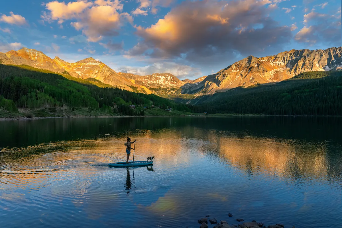 A serene lake scene featuring a person paddleboarding with their dog, framed by a dramatic mountain range. The water reflects the surrounding natural beauty, and the atmosphere is peaceful yet adventurous.