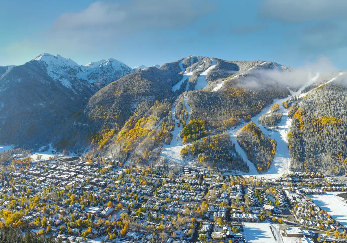 A panoramic view of snow-capped mountains and ski slopes, with vibrant autumn trees and a charming town nestled in the valley below. The environment is both adventurous and inviting, reflecting seasonal transitions.