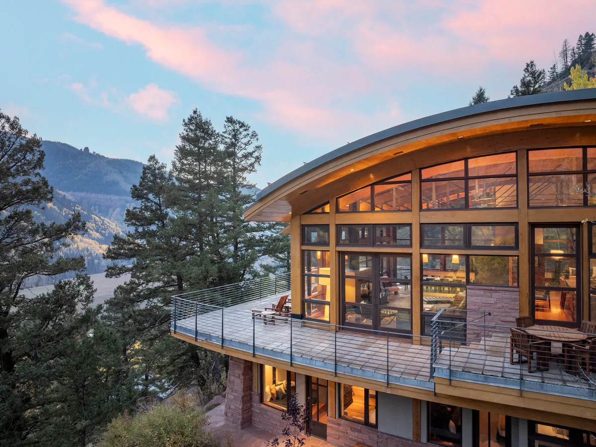 A wide deck area with comfortable seating is framed by a curved roof and large glass windows, opening onto lush trees and a stunning mountain view. The space is ideal for entertaining, relaxing at dusk, or stargazing, seamlessly connecting the home to its majestic surroundings.