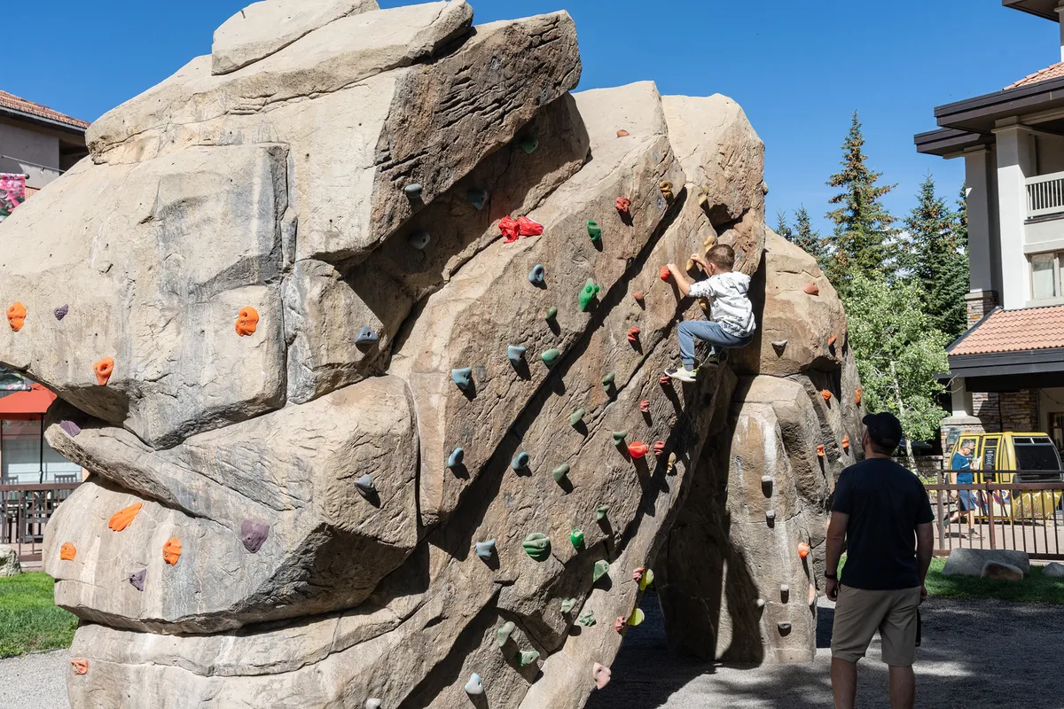 A child is actively climbing a rock wall as an adult supervises, set in an outdoor play environment. The focus is on encouraging adventure and safe, supervised play in a beautiful, natural setting.