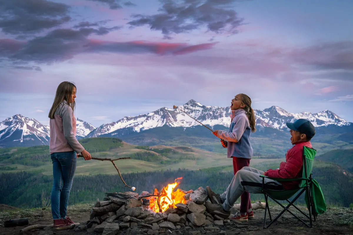 Children roast marshmallows around a campfire, with a parent nearby and mountains in the background. The scene radiates warmth and togetherness, capturing a quintessential family memory-making moment amid nature.
