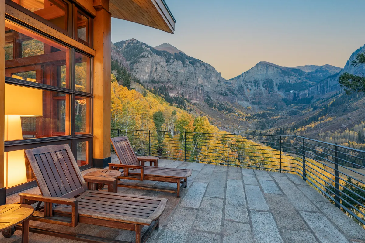 An expansive outdoor terrace with modern deck chairs is set against a breathtaking backdrop of the San Juan Mountains. Glass walls blend indoor and outdoor living, inviting relaxation and offering panoramic sunset views. The setting is serene, perfect for unwinding with a glass of wine and soaking in mountain beauty.