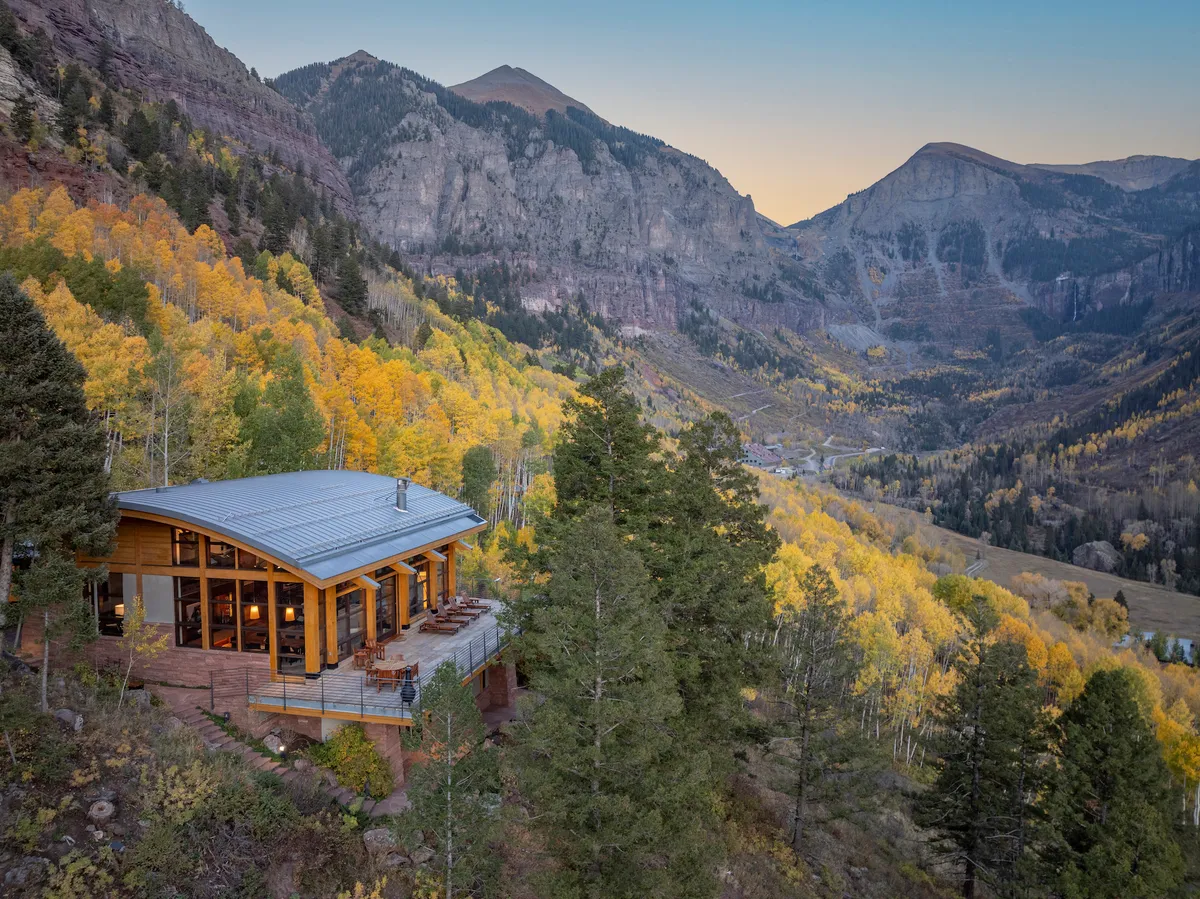 A sweeping exterior view of the modern Telluride retreat with vivid autumn foliage surrounding the house and dramatic mountain peaks in the background. The home’s clean lines and large windows emphasize its integration with the natural landscape, while the deck suggests opportunities for outdoor relaxation and enjoying the scenery.